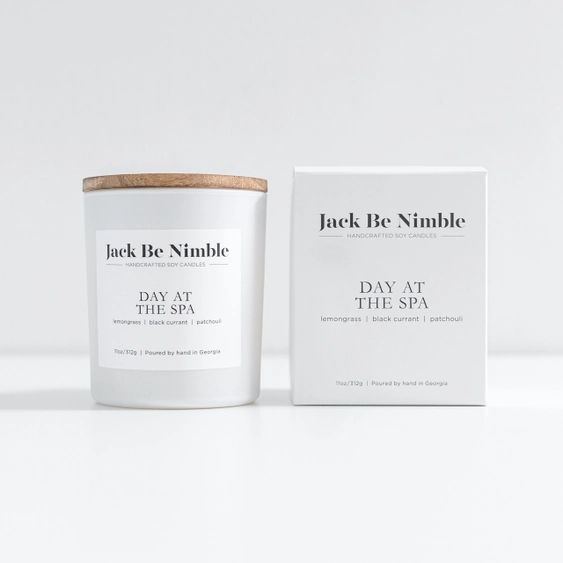A white jar candle with a light wood lid stands next to its white packaging box. Both the candle and box display the name &