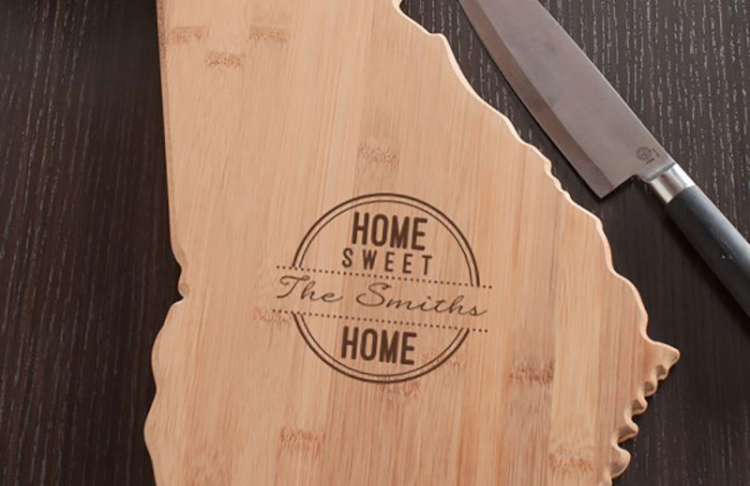 Personalized wooden cutting board shaped like a state of Georgia with 'Home Sweet The Smiths Home' engraving, accompanied by a knife on a dark surface.