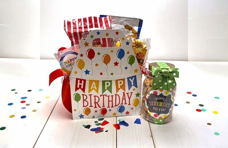Birthday gift box with colorful balloons and a jar of cookies and snacks on a white surface with confetti.