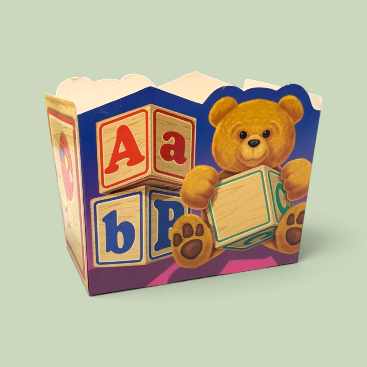 A decorative paper party favor box featuring a teddy bear holding a toy block and colorful alphabet blocks, set against a solid pale green background.