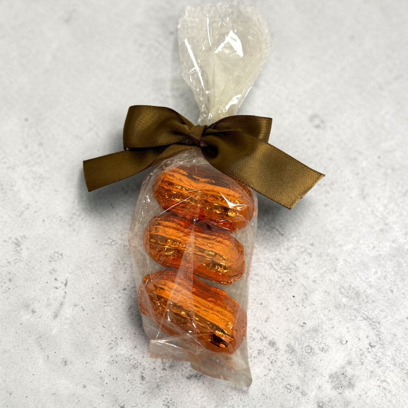 Three individually wrapped chocolate shaped peanuts filled with caramel tied with a brown satin ribbon in a clear cellophane bag, set against a light gray textured background.