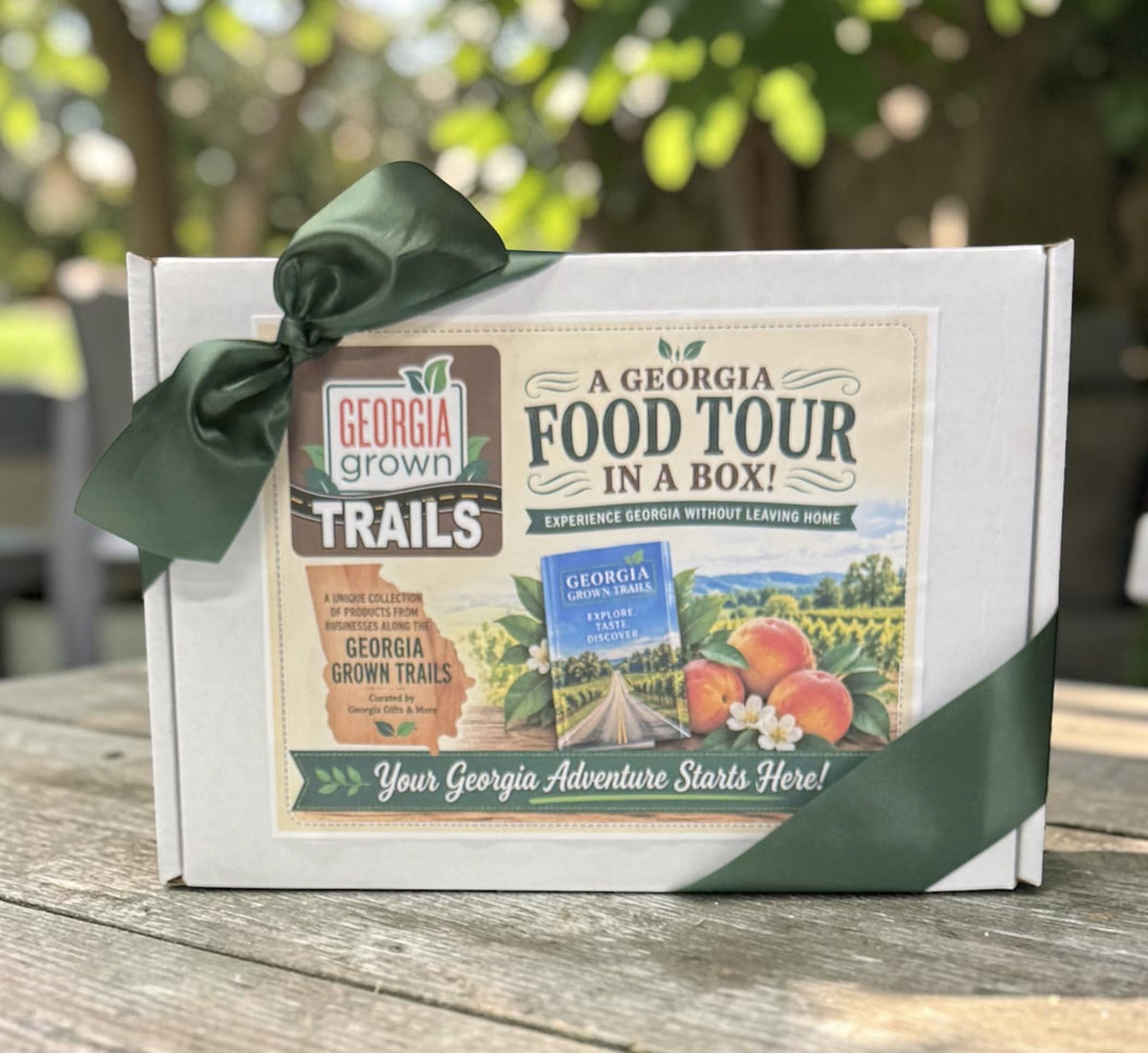 A white gift box tied with a dark green ribbon, featuring printed artwork for a Georgia Food Tour in a Box and Georgia Grown Trails, placed on a wooden table outdoors with blurred greenery in the background.