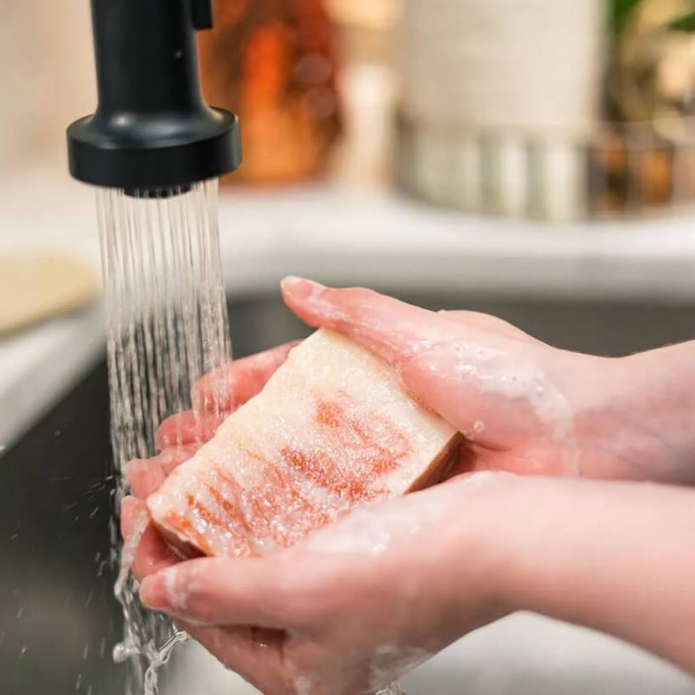 Person holding a piece of Georgia peach soap under running water in a kitchen sink.