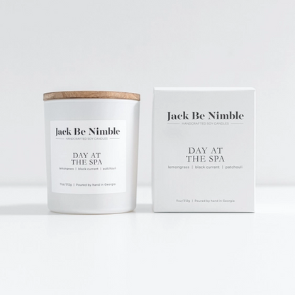 A white jar candle with a light wood lid stands next to its white packaging box. Both the candle and box display the name &
