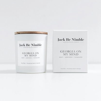 A peach scented soy candle jar with a light wood lid sits beside its white product box. The labels read Georgia On My Mind, with small scent notes peach, watermelon, and honeysuckle.