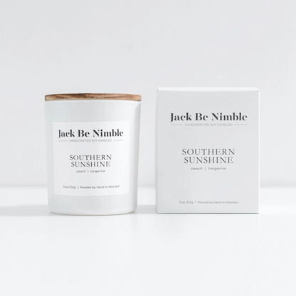 A scented soy candle in a white jar with a light wood lid sits beside its matching white box. The label reads Southern Sunshine, with small peach and tangerine text and a &