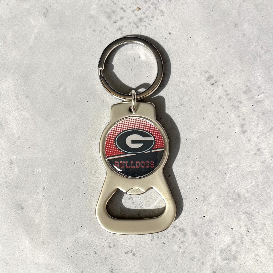 A metal bottle opener keychain featuring the Georgia Bulldogs logo, resting on a light gray textured surface.