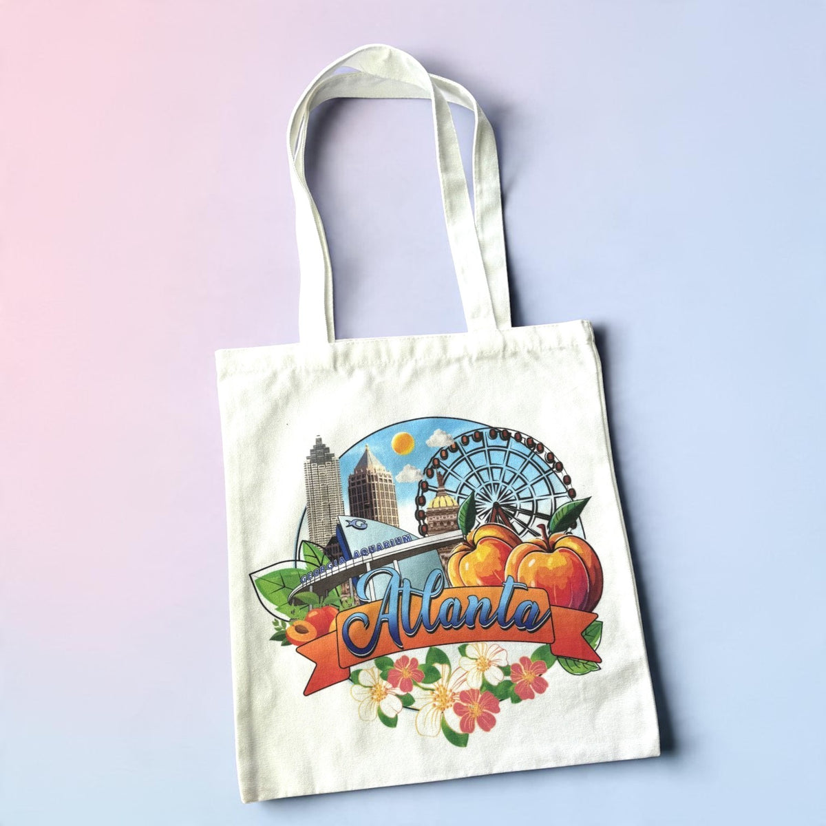 A cream-colored canvas tote bag featuring a colorful graphic illustration of Atlanta, Georgia, including the skyline, Ferris wheel, peaches, flowers, and the word &