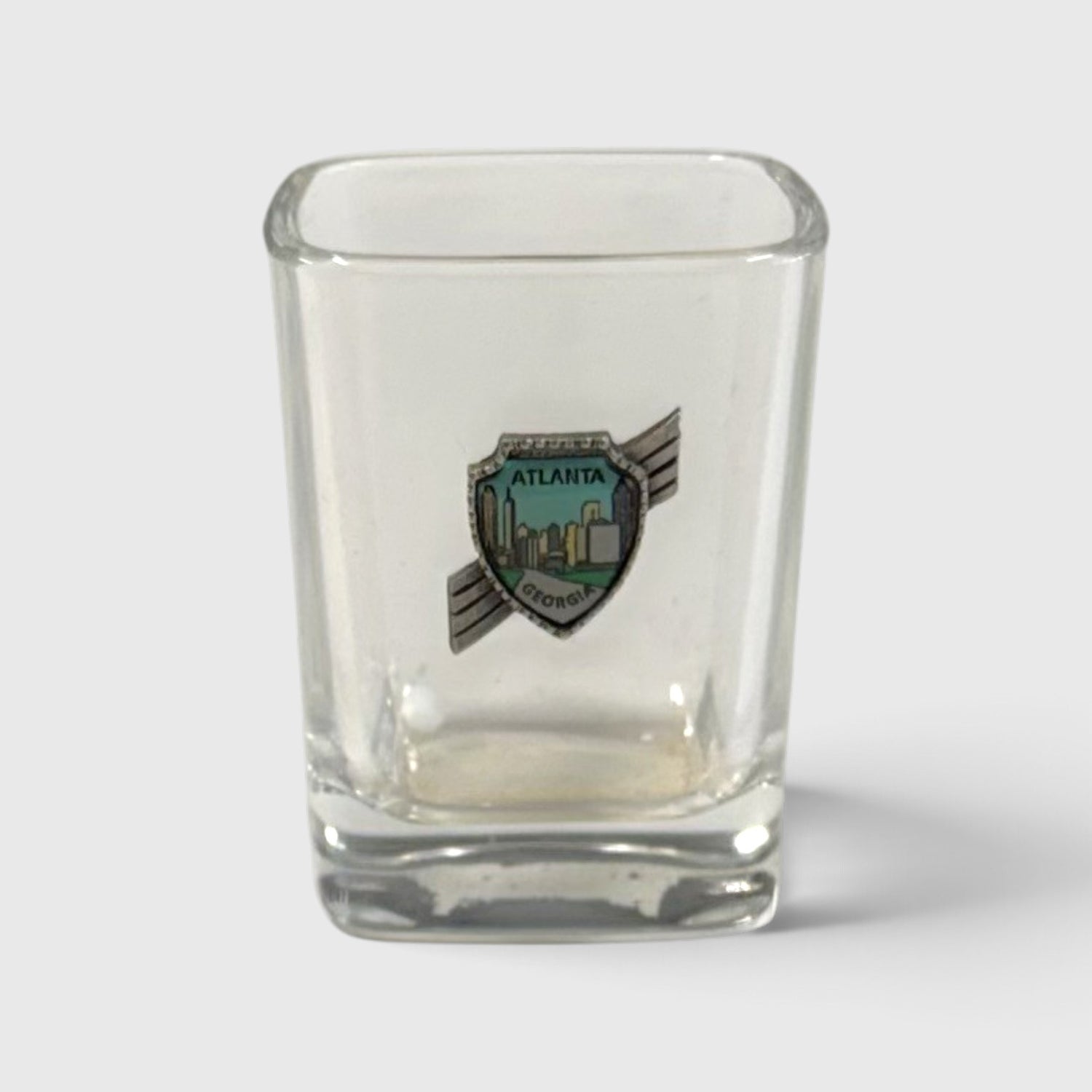 A clear square glass shot glass featuring a shield-shaped Atlanta city emblem on the front, set against a light gray background.