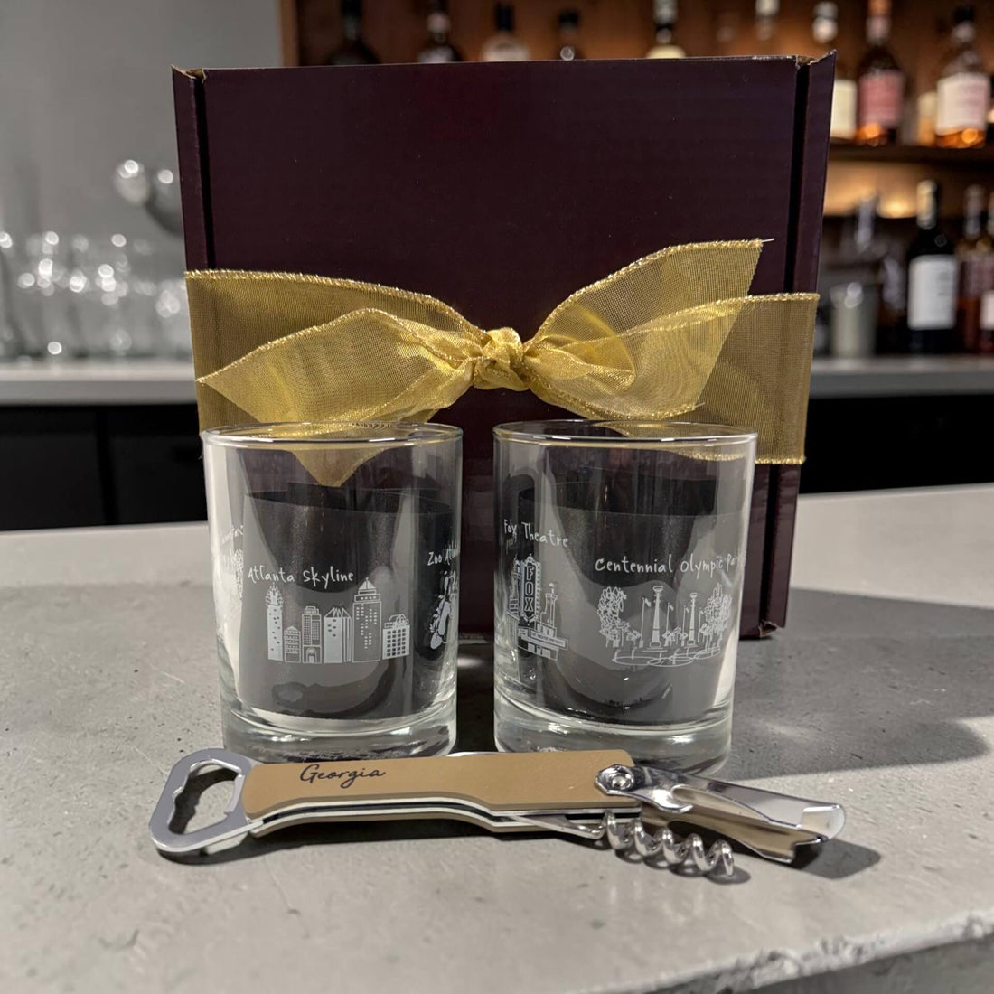 Two shot glasses with city skyline etching, a gold ribbon, and a bottle opener on a counter.