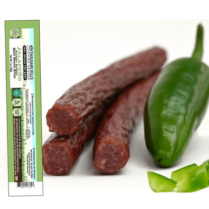 Two dried jalapeno pepper beef sticks with a green label on the left and a fresh jalapeno pepper on the right.