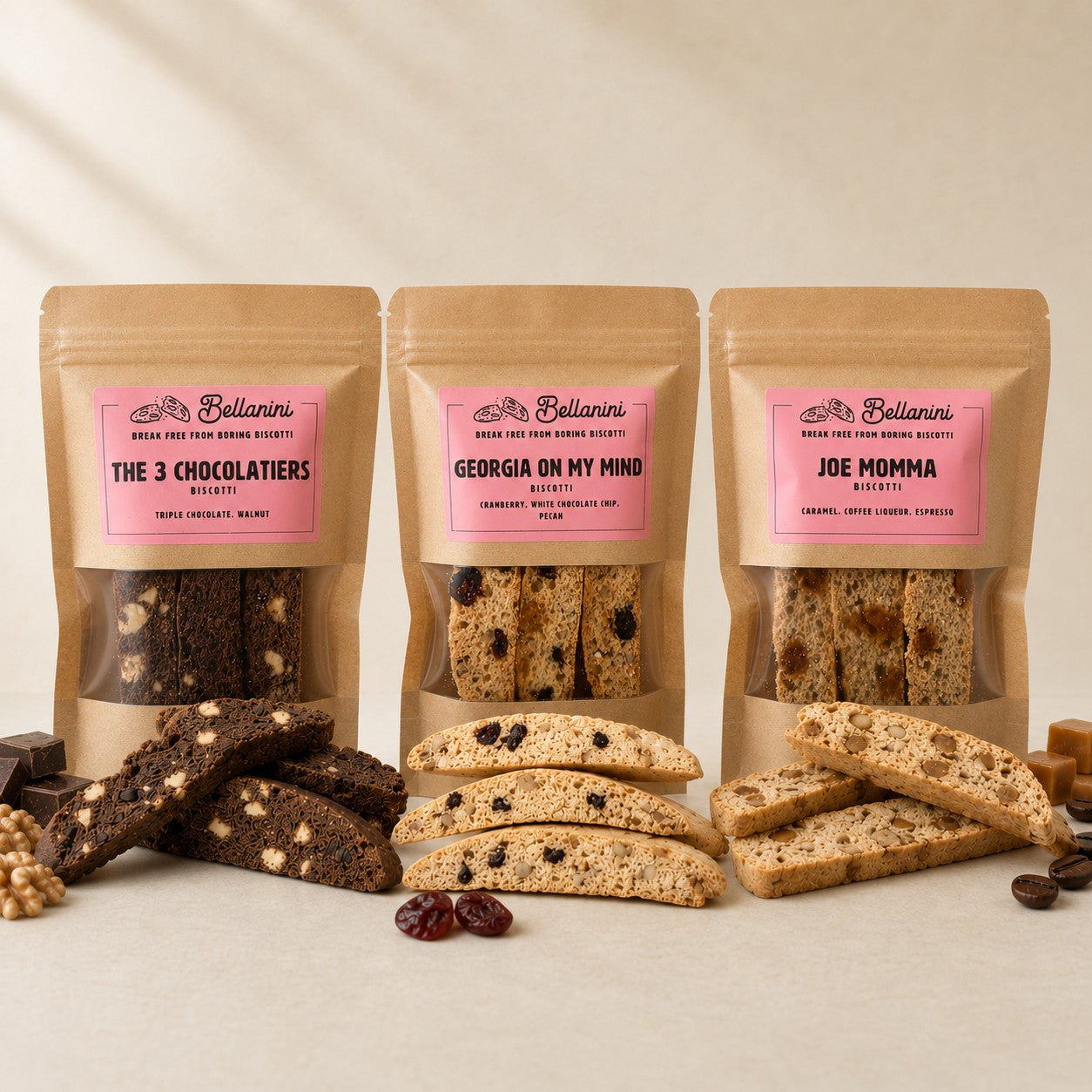Three stand-up pouches of Bellanini biscotti cookies arranged on a light beige surface with loose biscotti pieces and dried fruit in front. The center pouch is labeled Georgia On My Mind, flanked by The 3 Chocolatiers on the left and Joe Momma on the right.