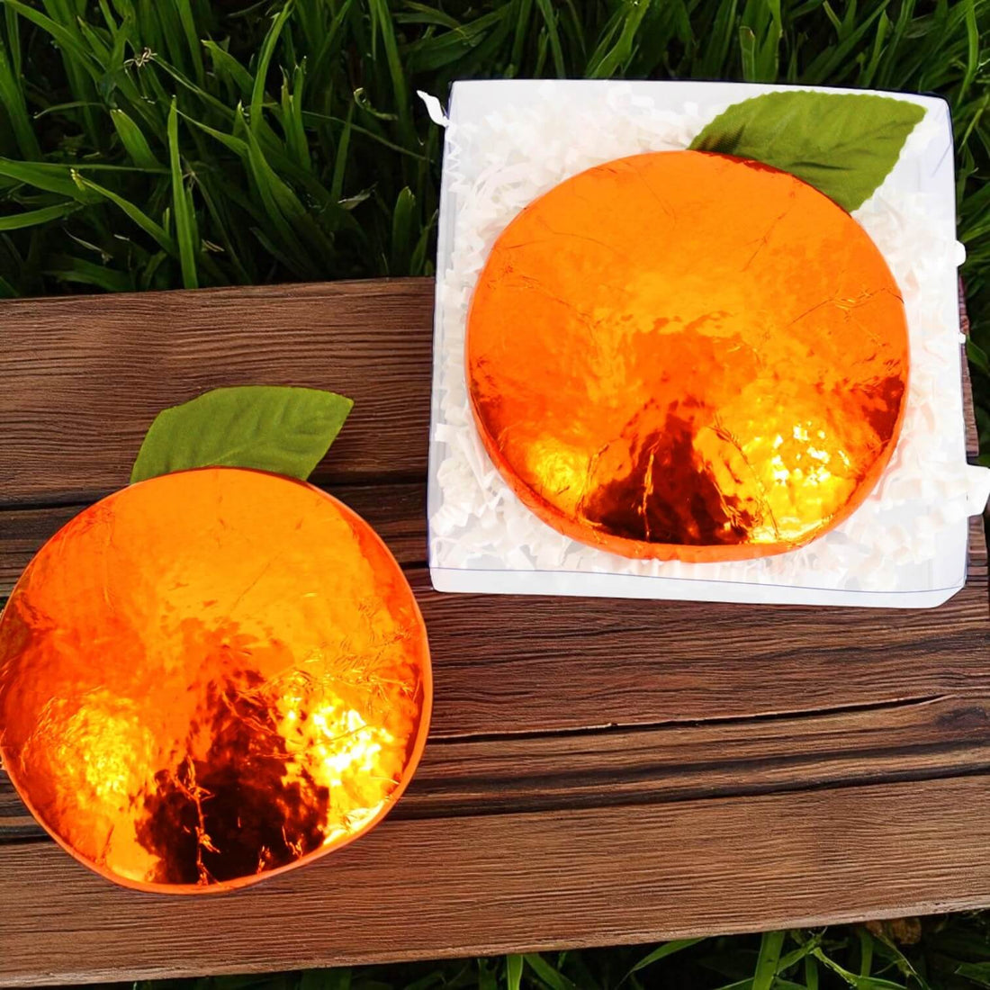 Two foil wrapped chocolates in the shape of oranges on a wooden surface with grass in the background.