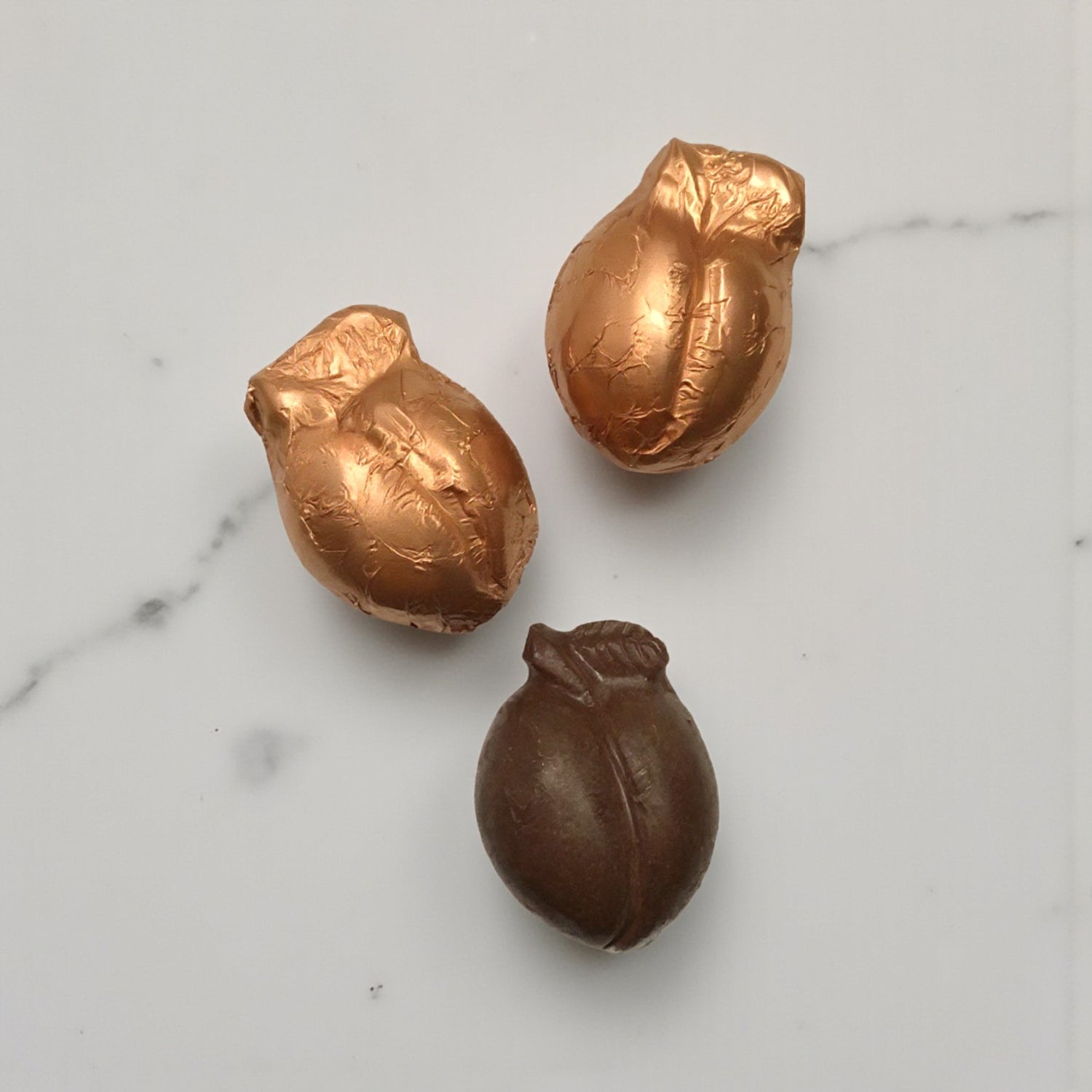 Three peach-shaped chocolate candies with copper foil wrappers on a marble surface