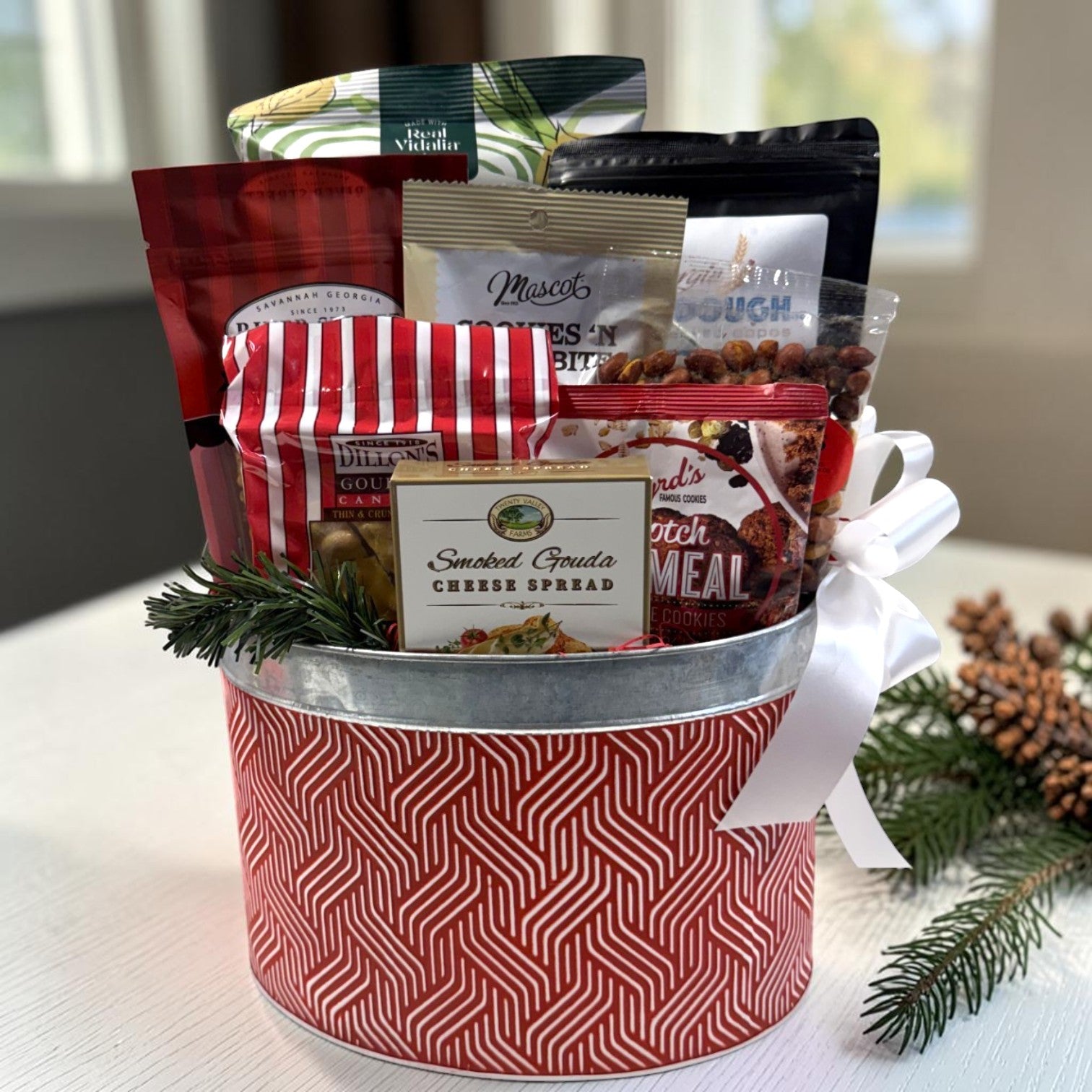A holiday gift basket filled with assorted gourmet snacks and treats, including Peanut Brittle, Popcorn, Chocolates, Nuts, Cookies and Cheese Spread, wrapped in a red and white patterned tin with a white ribbon bow.