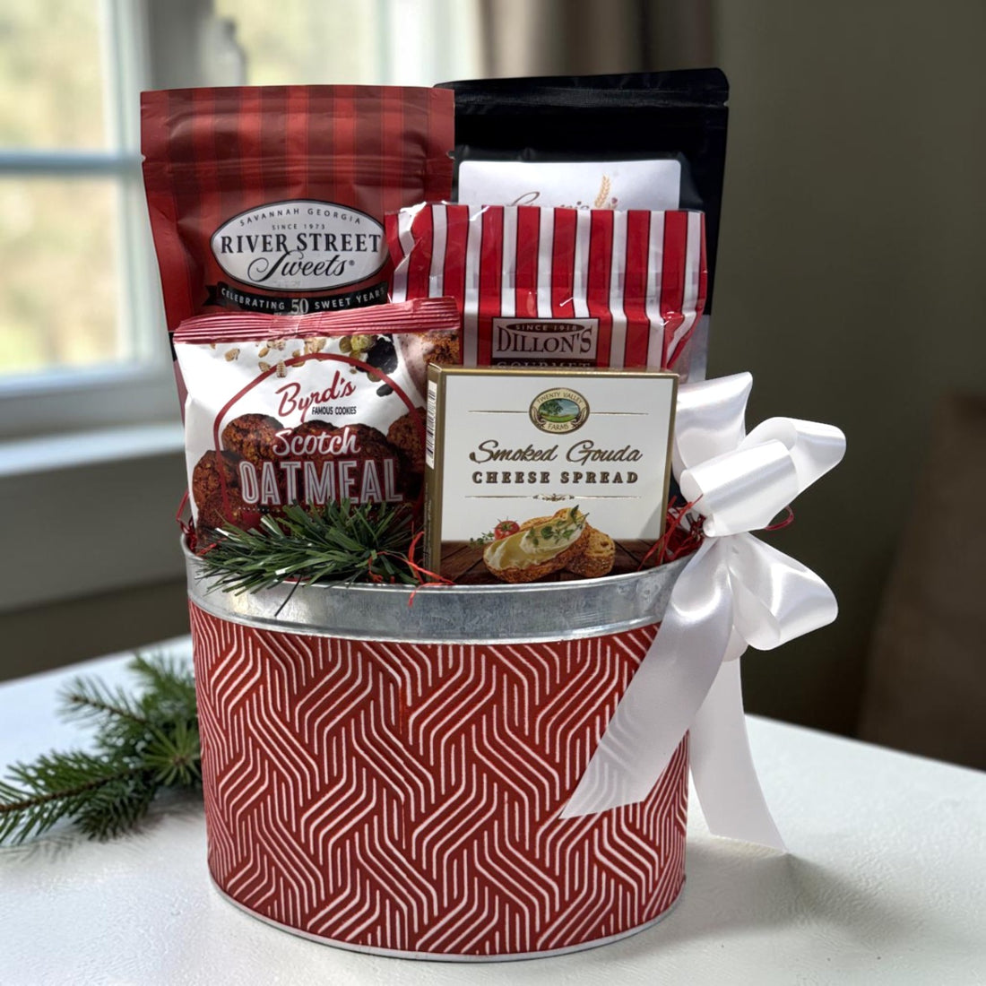 A holiday-themed gift basket filled with assorted gourmet snacks and spreads, wrapped in a red and white patterned tin with a white ribbon bow. Visible items include River Street Sweets, Byrd&