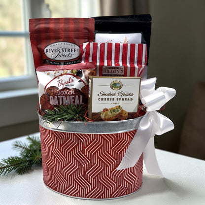 A holiday-themed gift basket filled with assorted gourmet snacks and spreads, wrapped in a red and white patterned tin with a white ribbon bow. Visible items include River Street Sweets, Byrd&