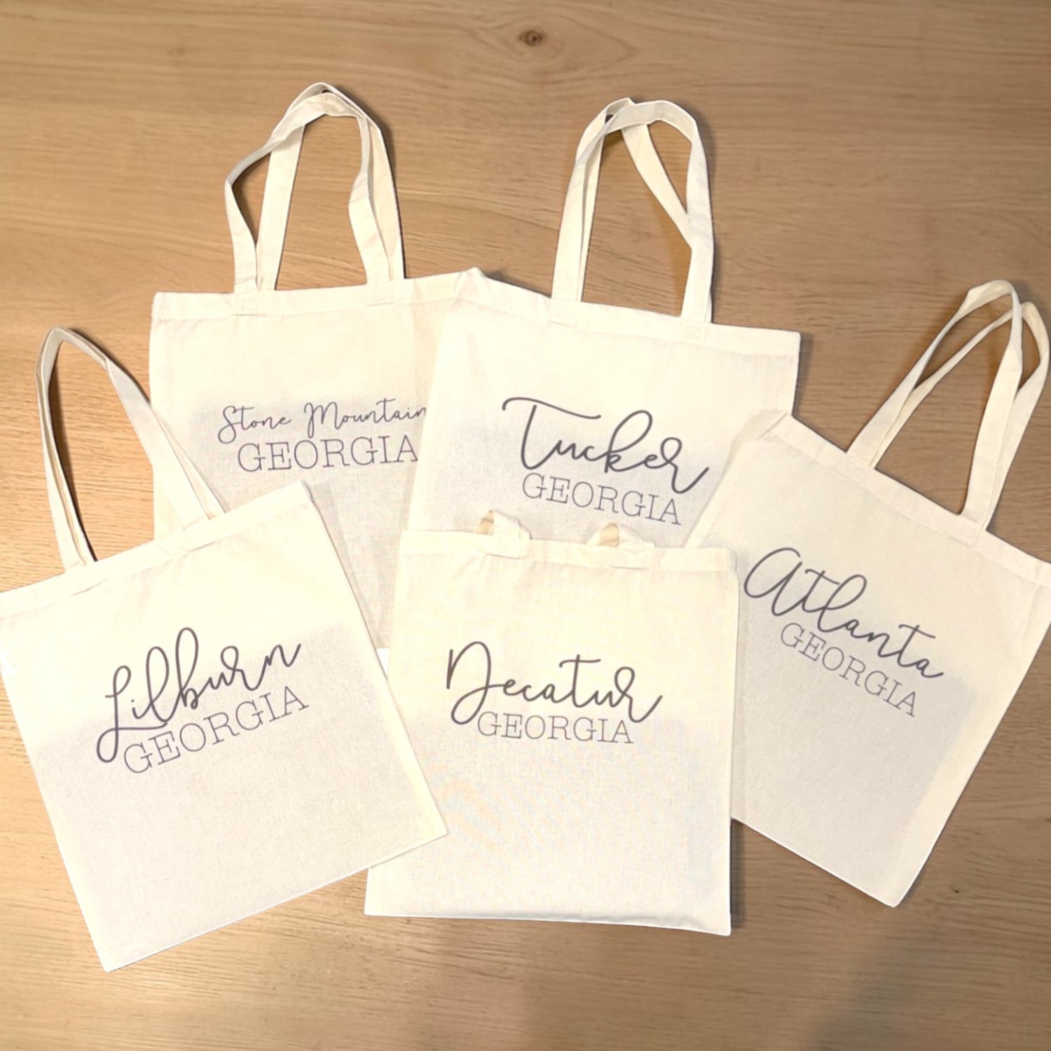 Five canvas tote bags with various city names printed on them on a wooden surface