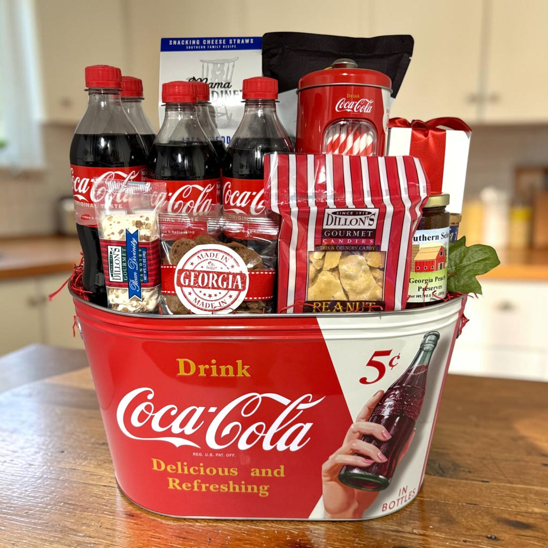 A Coca-Cola-themed gift basket arranged in a red metal tub on a wooden table, with several Coca-Cola bottles, cookies, divinity, snacks, and peach preserves shown in a kitchen setting.
