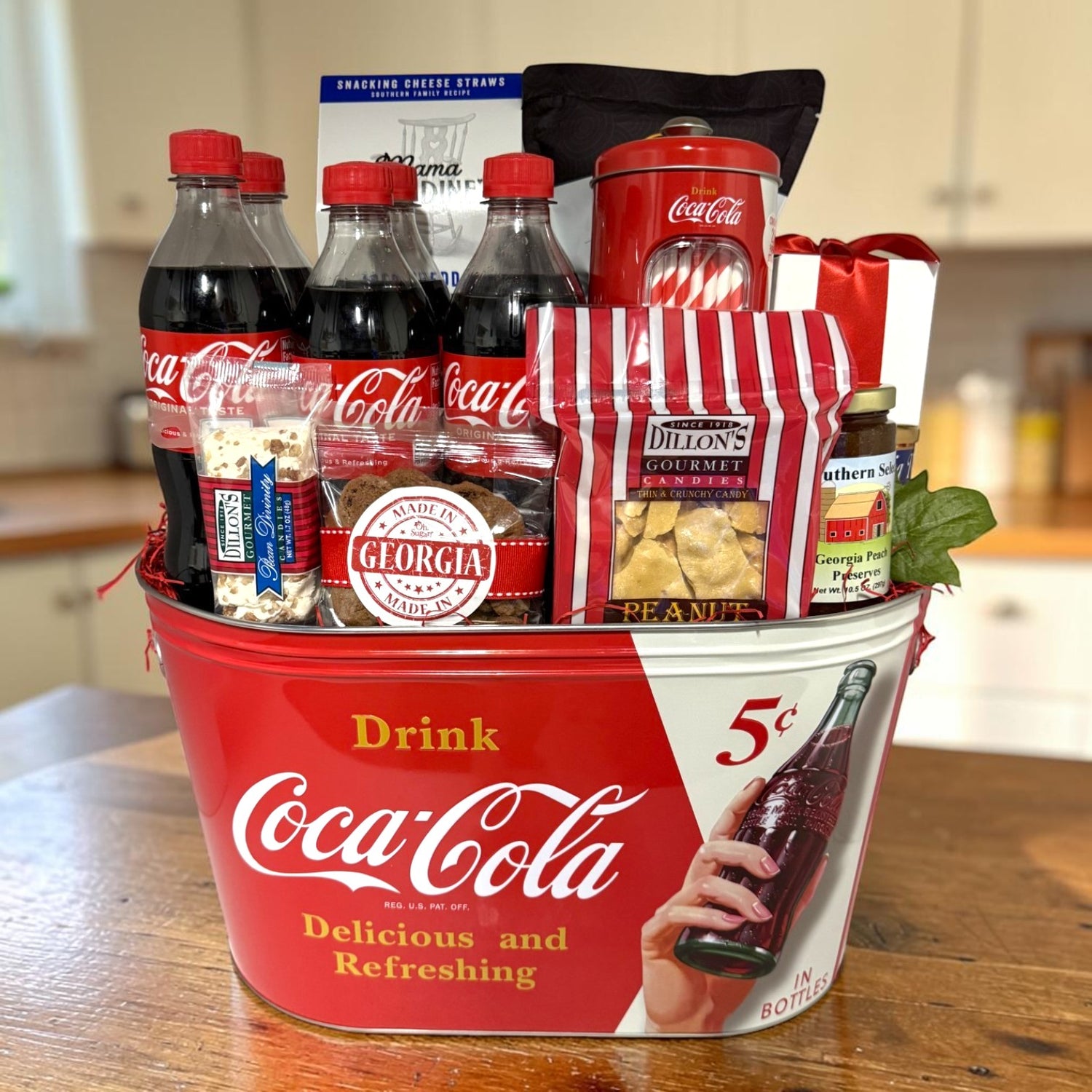 A Coca-Cola-themed gift basket arranged in a red metal tub on a wooden table, with several Coca-Cola bottles, cookies, divinity, snacks, and peach preserves shown in a kitchen setting.
