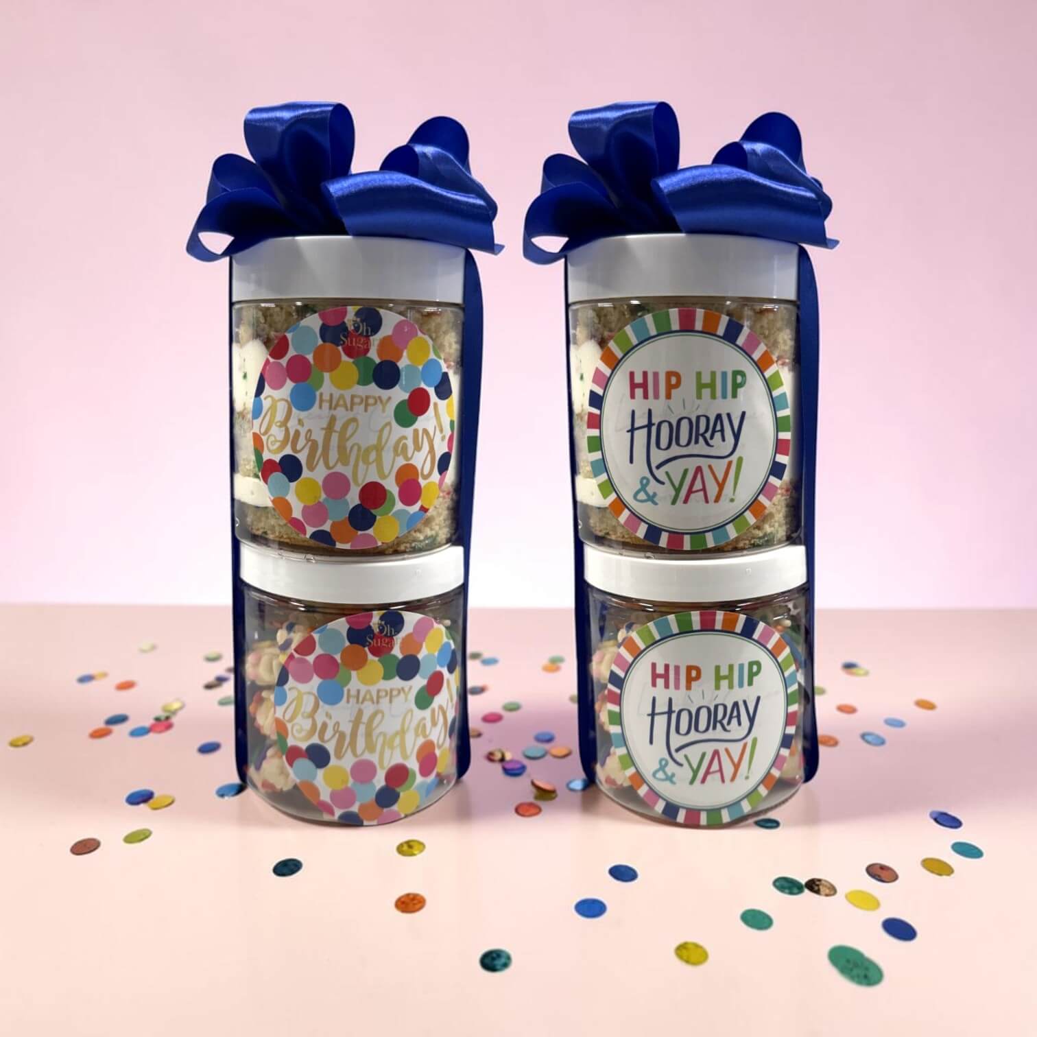 Two stacks of cake and cookie combos with blue ribbons and celebratory labels on a pink background with confetti.