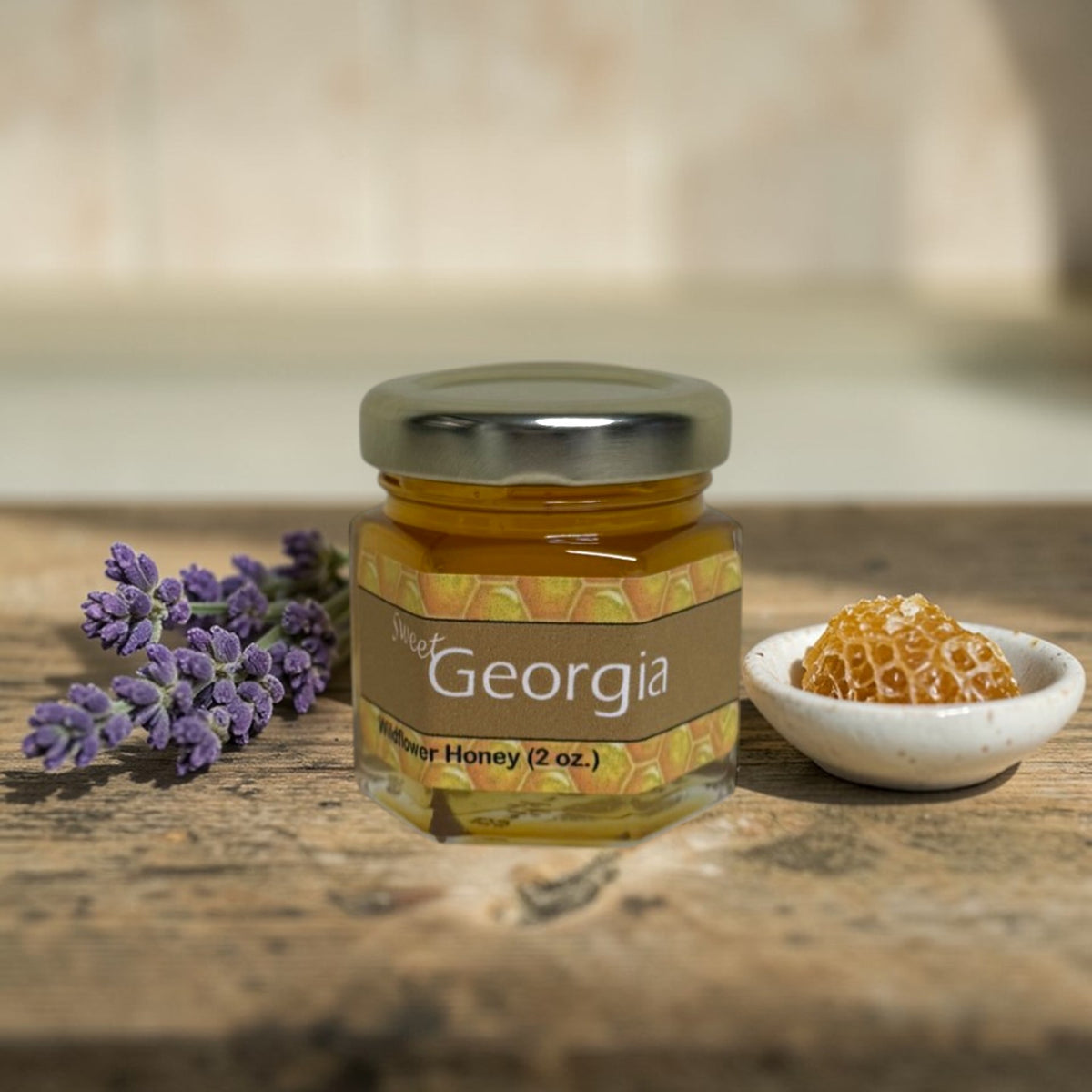 A small glass jar of Sweet Georgia Wildflower Honey sits on a rustic wooden surface, flanked by a sprig of purple lavender on the left and a small white dish of honeycomb on the right. The background is a soft-focus beige wall.