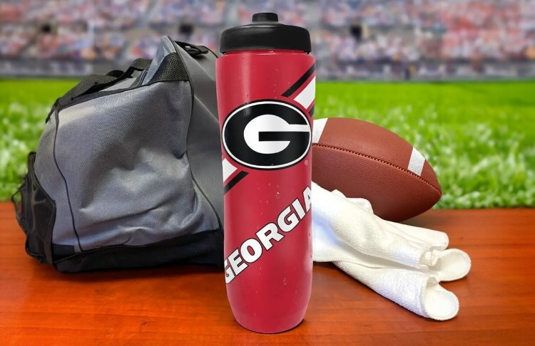 Red water bottle with 'Georgia' logo, football, and sports bag on a table with stadium background