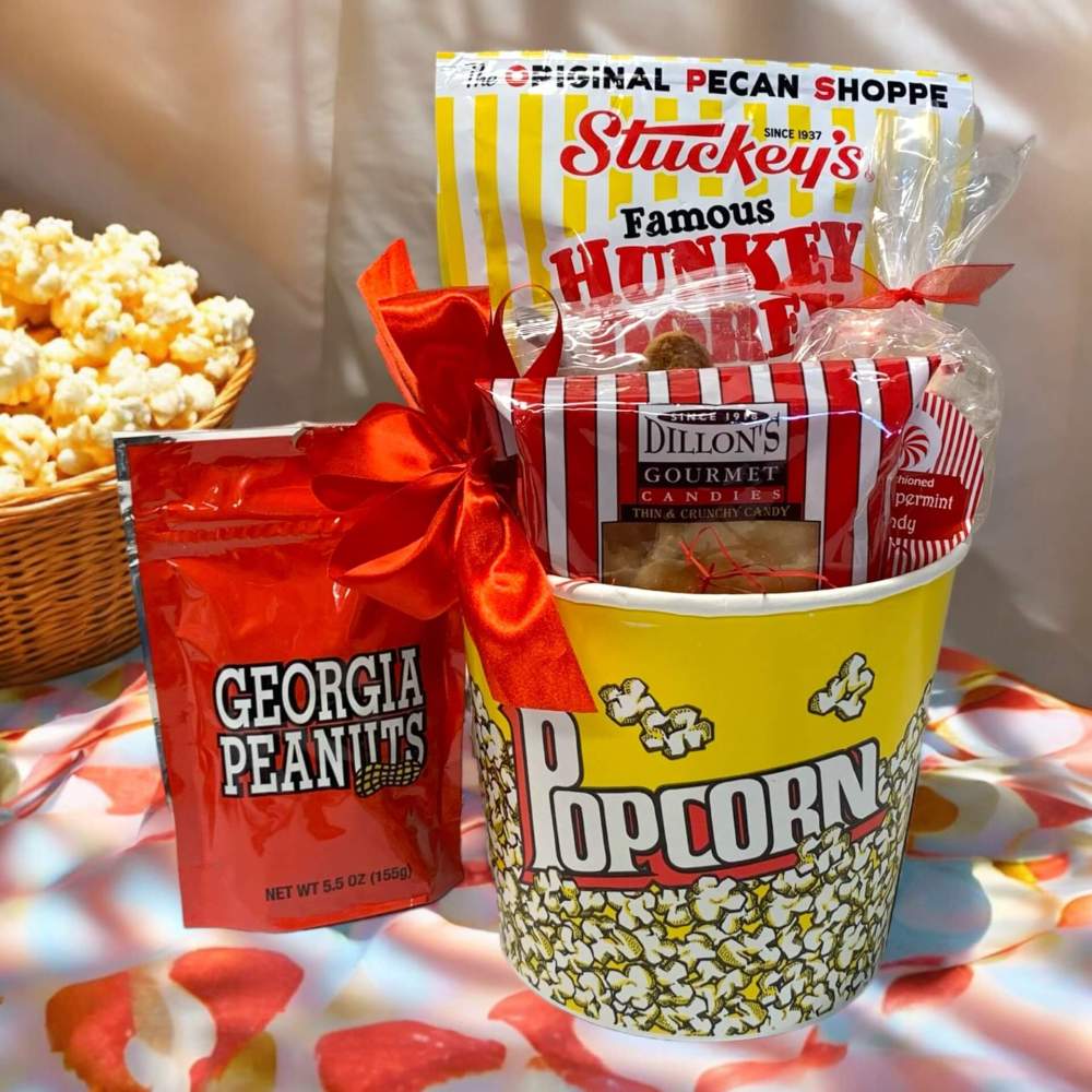 Paper popcorn bucket with gourmet popcorn, cookies, nuts and snacks with a bowl of popcorn in the background.