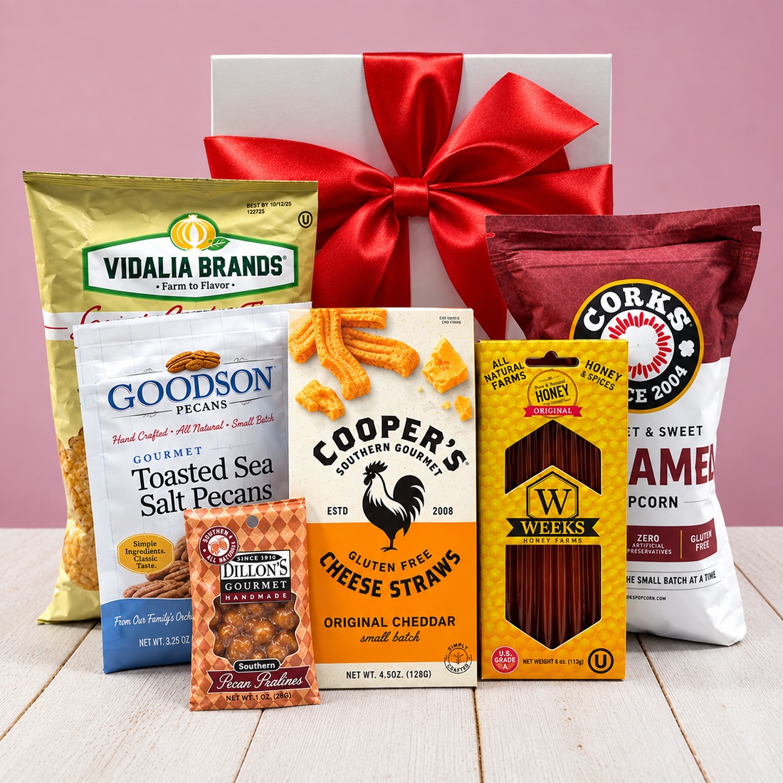 A gift box arrangement of gluten free snack and candy products on a light wood surface against a pink background, with a white box and large red bow behind the items. Visible brands include Vidalia Brands, Goodson Pecans, Cooper&