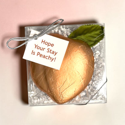 Gold peach-shaped chocolate in clear gift box with a Hope Your Stay Is Peachy tag on a pink background