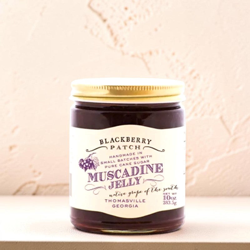 A jar of Muscadine Jelly by Blackberry Patch, featuring a cream-colored label with purple text and a gold metal lid, set against a textured beige background.