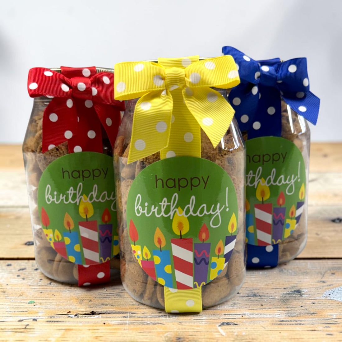 Three clear jars filled with cookies, tied with colorful polka-dot ribbons and decorated with round green labels that say "happy birthday!" with candle illustrations, displayed on a rustic wooden surface.