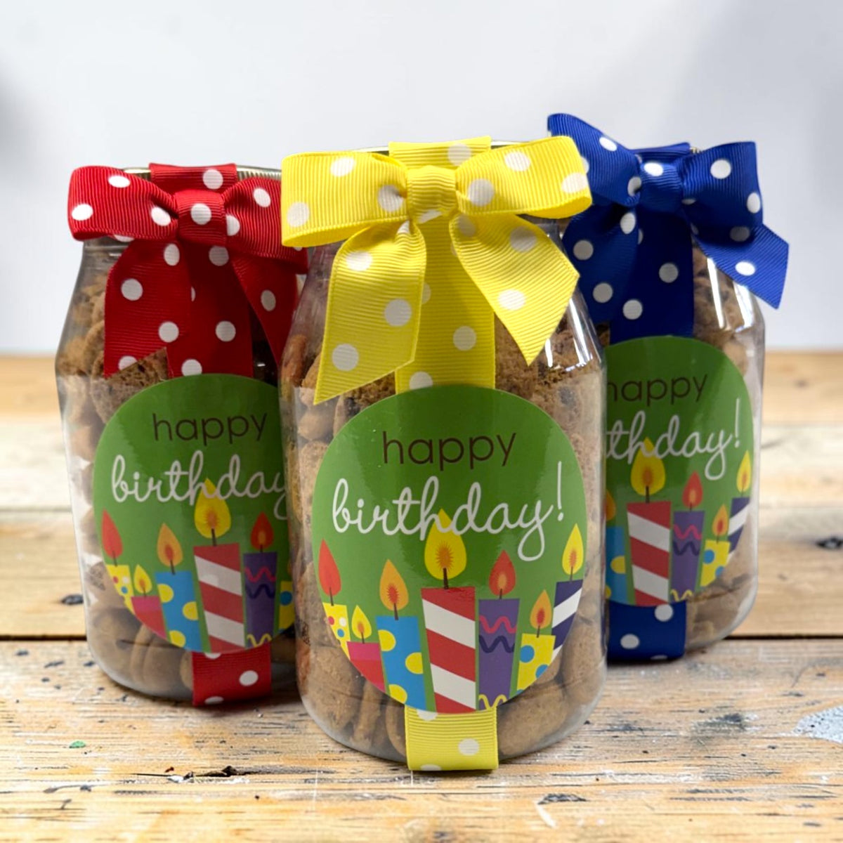 Three clear jars filled with cookies, tied with colorful polka-dot ribbons and decorated with round green labels that say "happy birthday!" with candle illustrations, displayed on a rustic wooden surface.