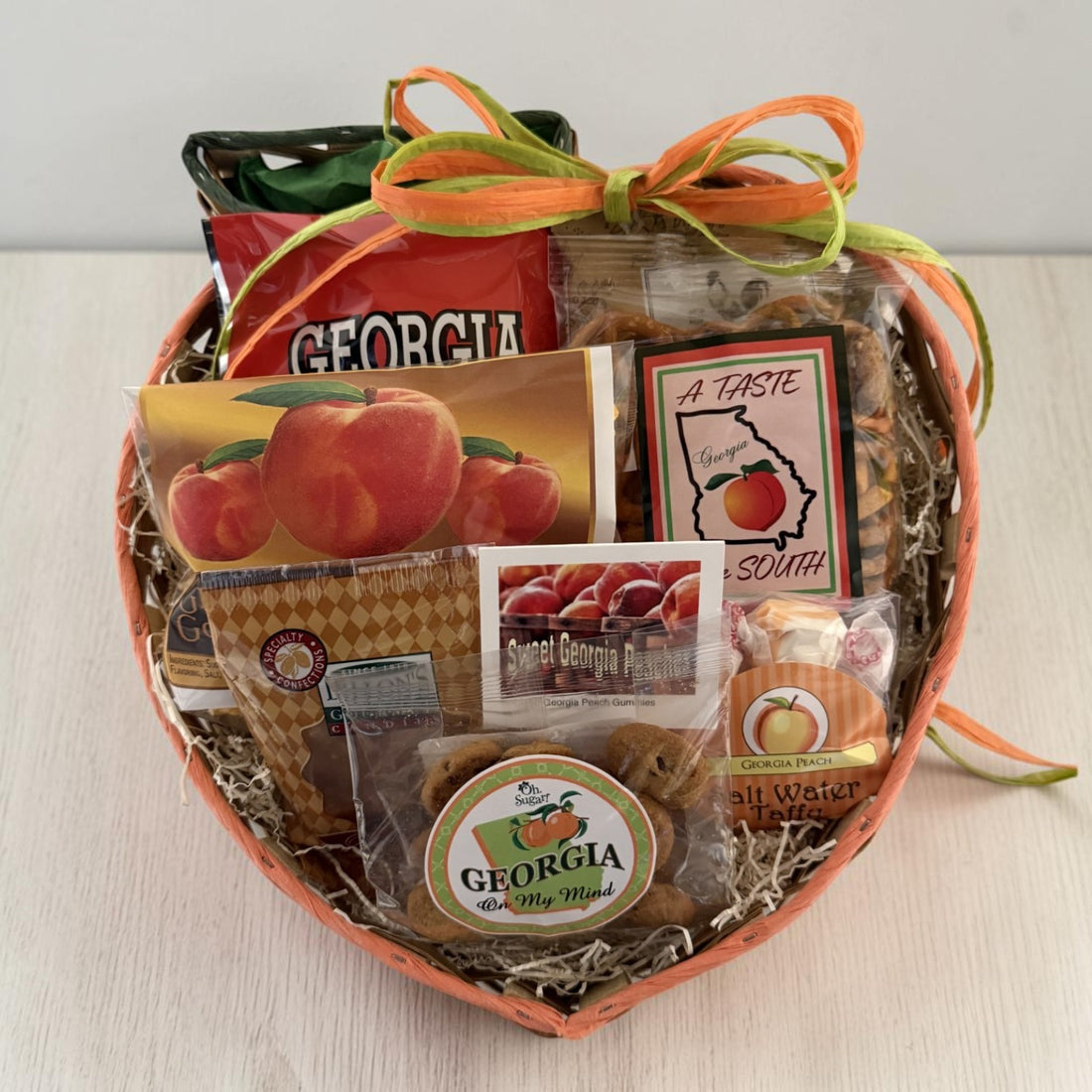 A peach-shaped gift basket filled with assorted Georgia-themed snacks, cookies, and packaged treats, tied with orange and green ribbons. Visible items include a bag of Georgia peach popcorn, cookies, snack mix, pralines and Georgia peach taffy on a light tabletop.