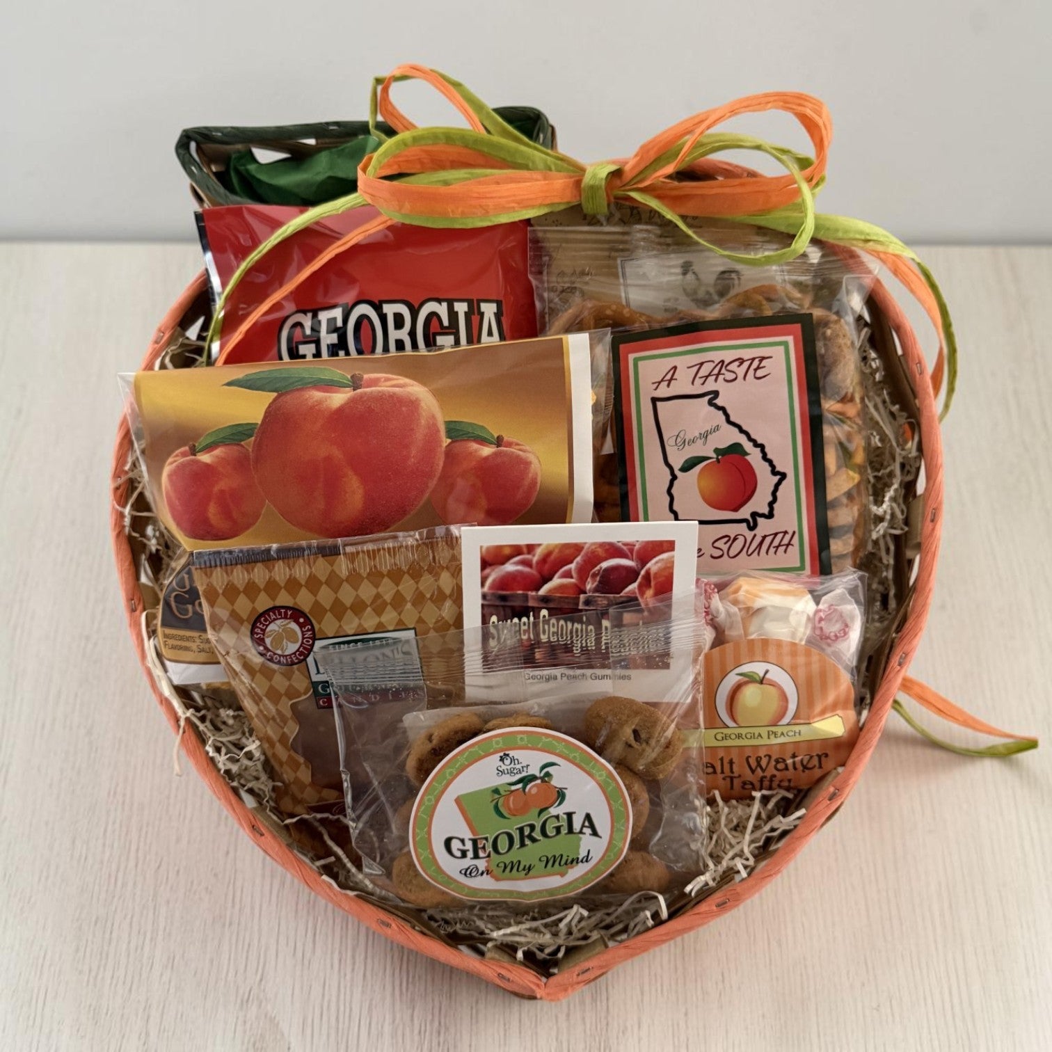 A peach-shaped gift basket filled with assorted Georgia-themed snacks, cookies, and packaged treats, tied with orange and green ribbons. Visible items include a bag of Georgia peach popcorn, cookies, snack mix, pralines and Georgia peach taffy on a light tabletop.