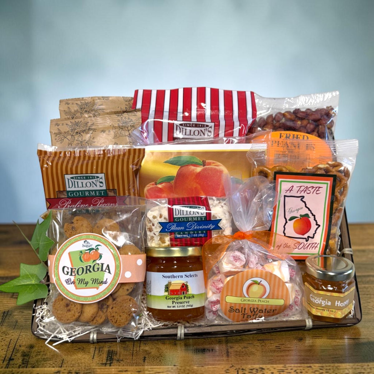 A gourmet gift basket filled with assorted Southern-style snacks and treats, including cookies, pretzels, peanut brittle, candy, peanuts, pecans, peach preserves and honey, arranged on a wooden tray over a table against a soft blue background.