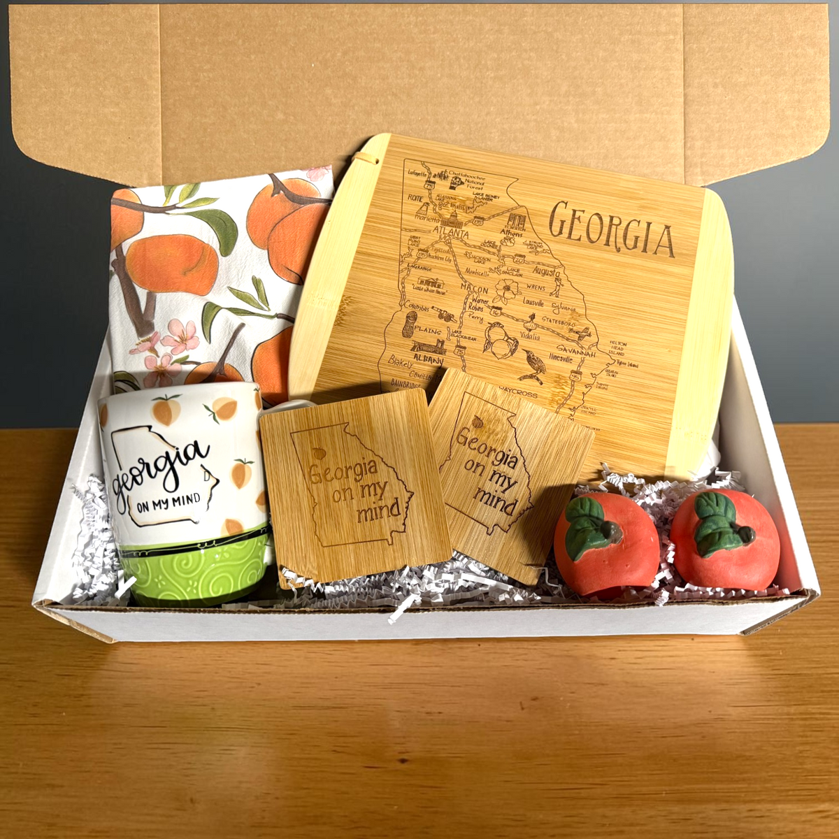 A curated gift box filled with Georgia-themed items, including an engraved bamboo cutting board, a ceramic mug, a patterned napkin, two small wooden coasters, and two peach shaped napkin rings, arranged in a white box on a wooden table.