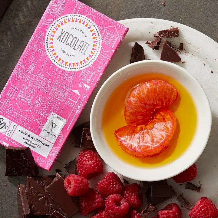 Pink Xocolati chocolate bar with a bowl of orange slices in syrup, raspberries, and chocolate pieces on a plate.